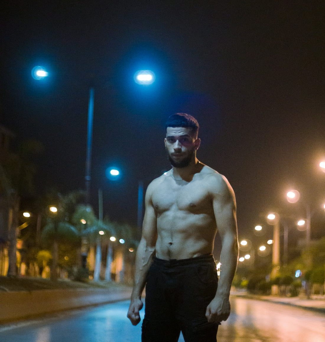 topless man in black pants wearing black sunglasses standing on sidewalk during night time